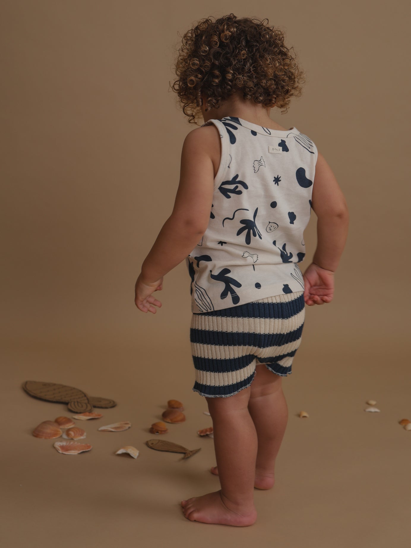 back view of toddler in capri white summer vest with navy print & striped shorts looking at seashells on floor