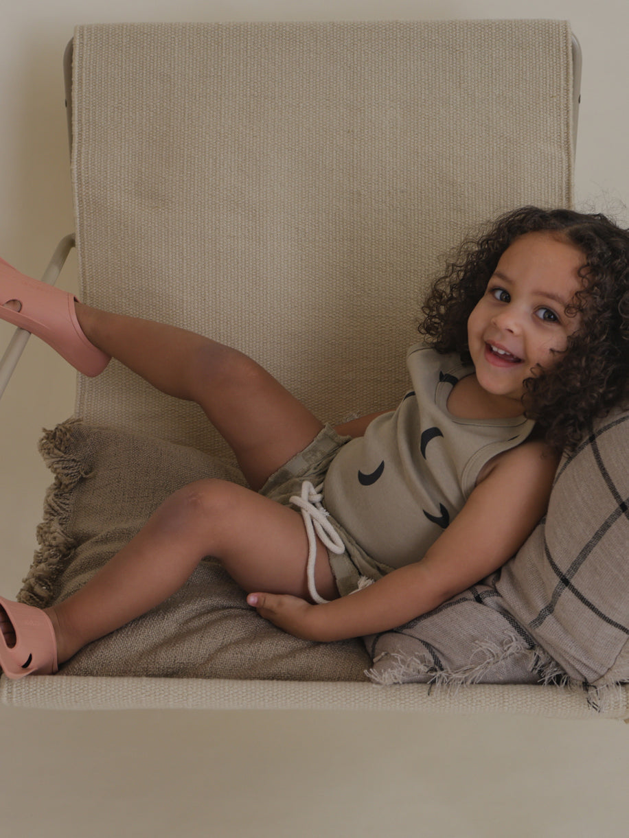 happy toddler in ribbed cotton beige vest with black moon print sitting sideways on chair
