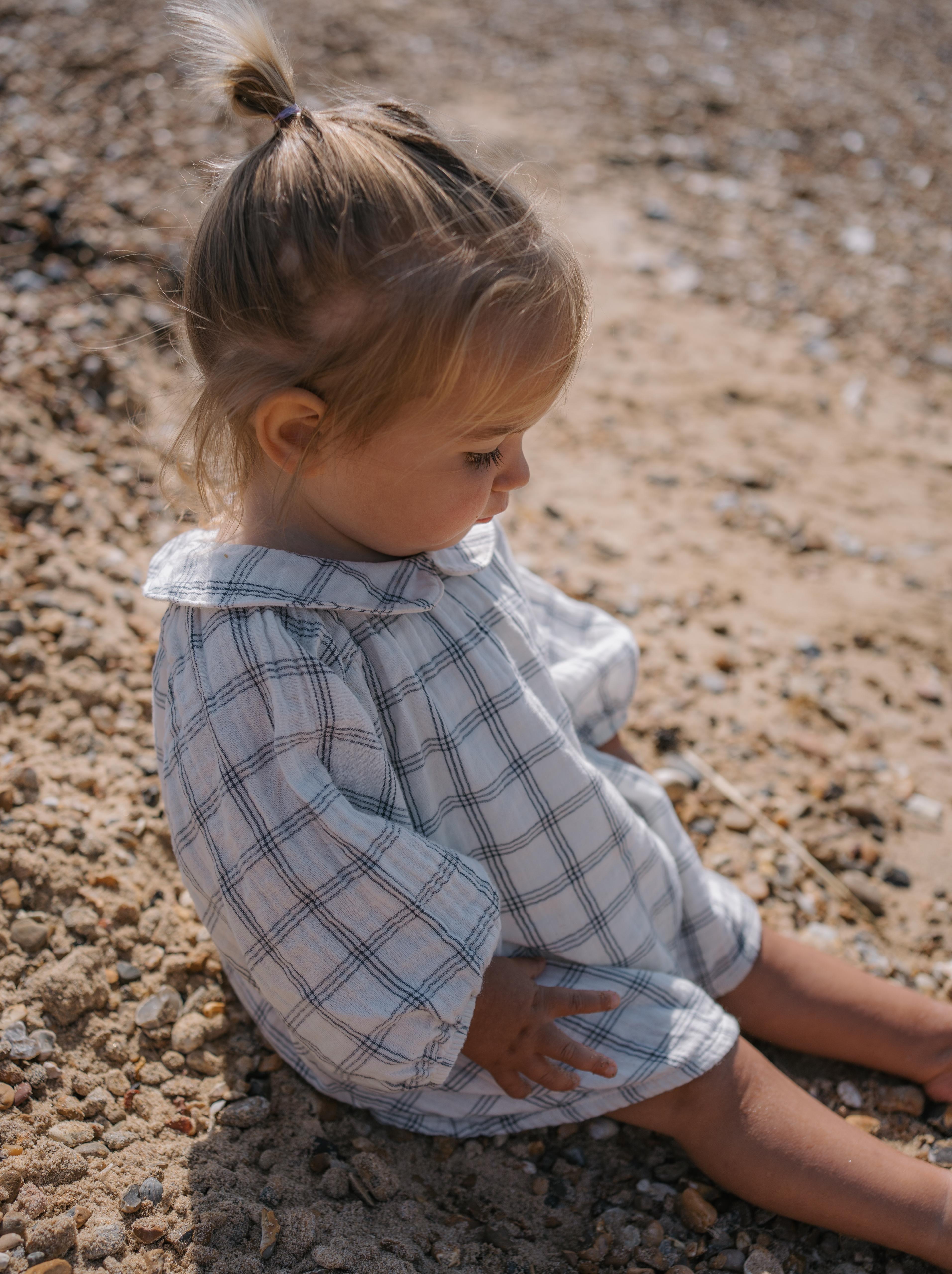 girl sitting on the beach wearing navy window check long sleeved two-way muslin button-down white dress, with peter pan collar & gathered cuffs