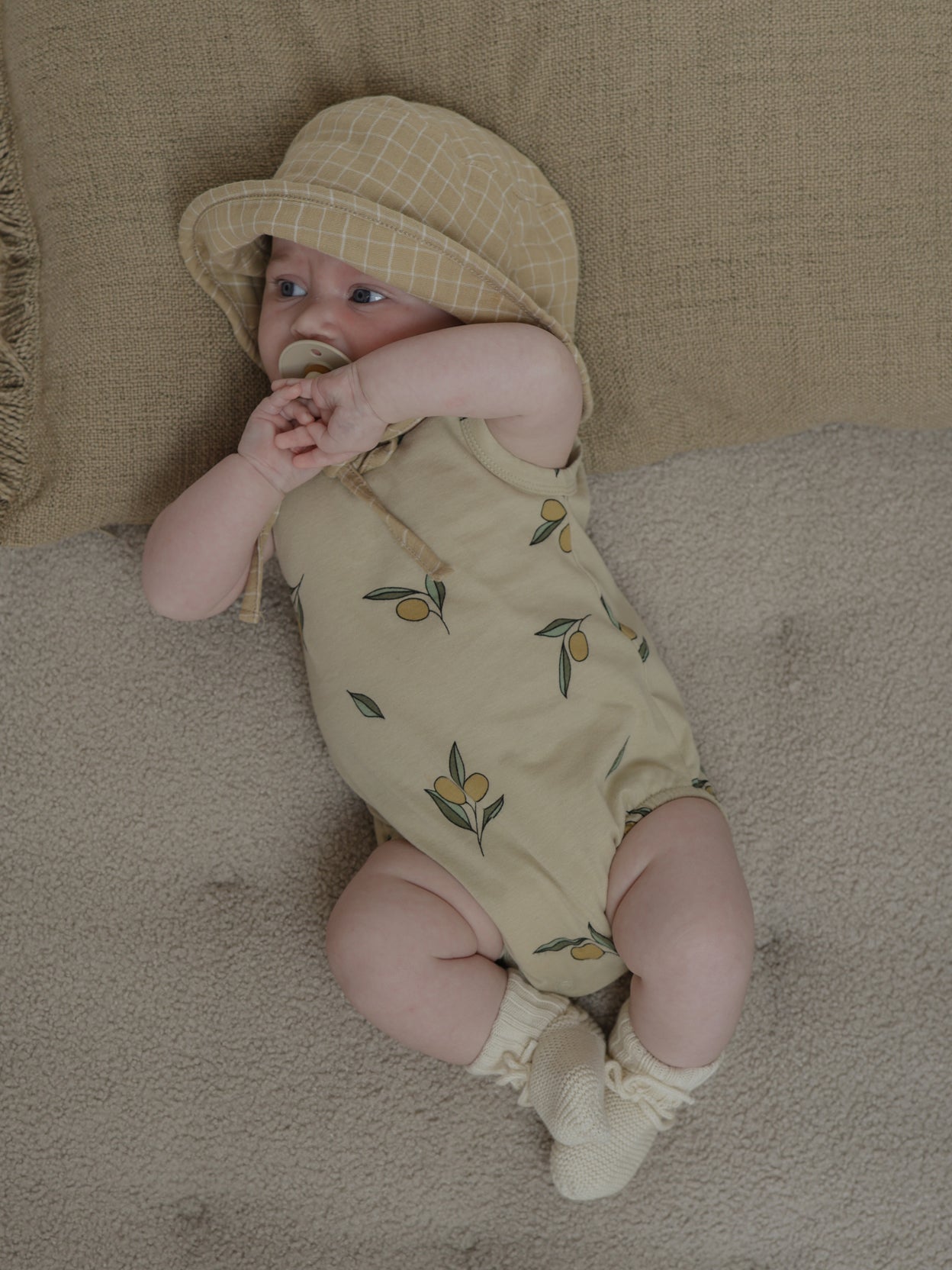 baby wearing cream sleeveless bodysuit with olives, hat & booties lying on cushion with hands together