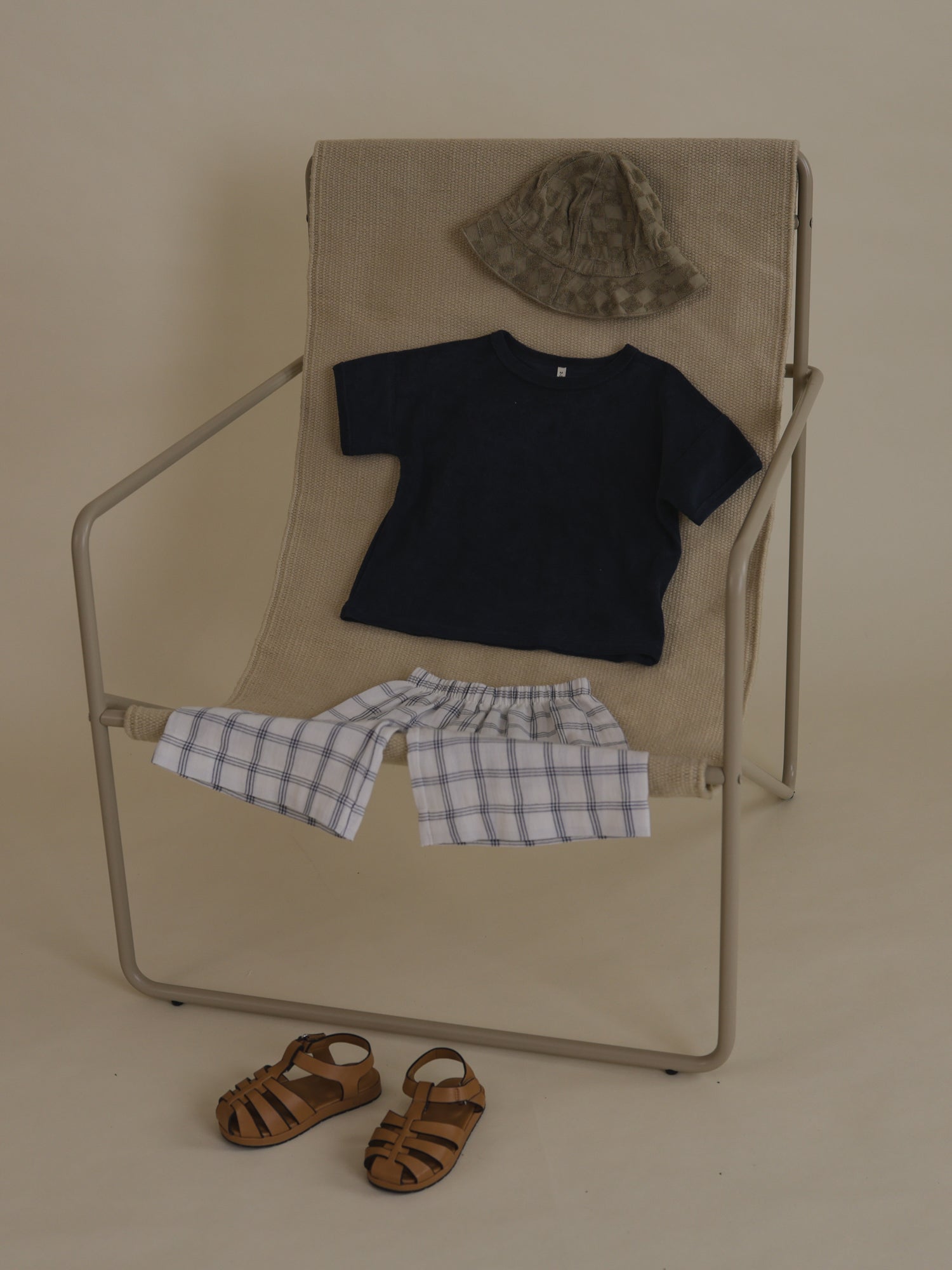 flat lay of navy terry t-shirt, grid pants, sun hat outfit on a chair, with sandals on the floor