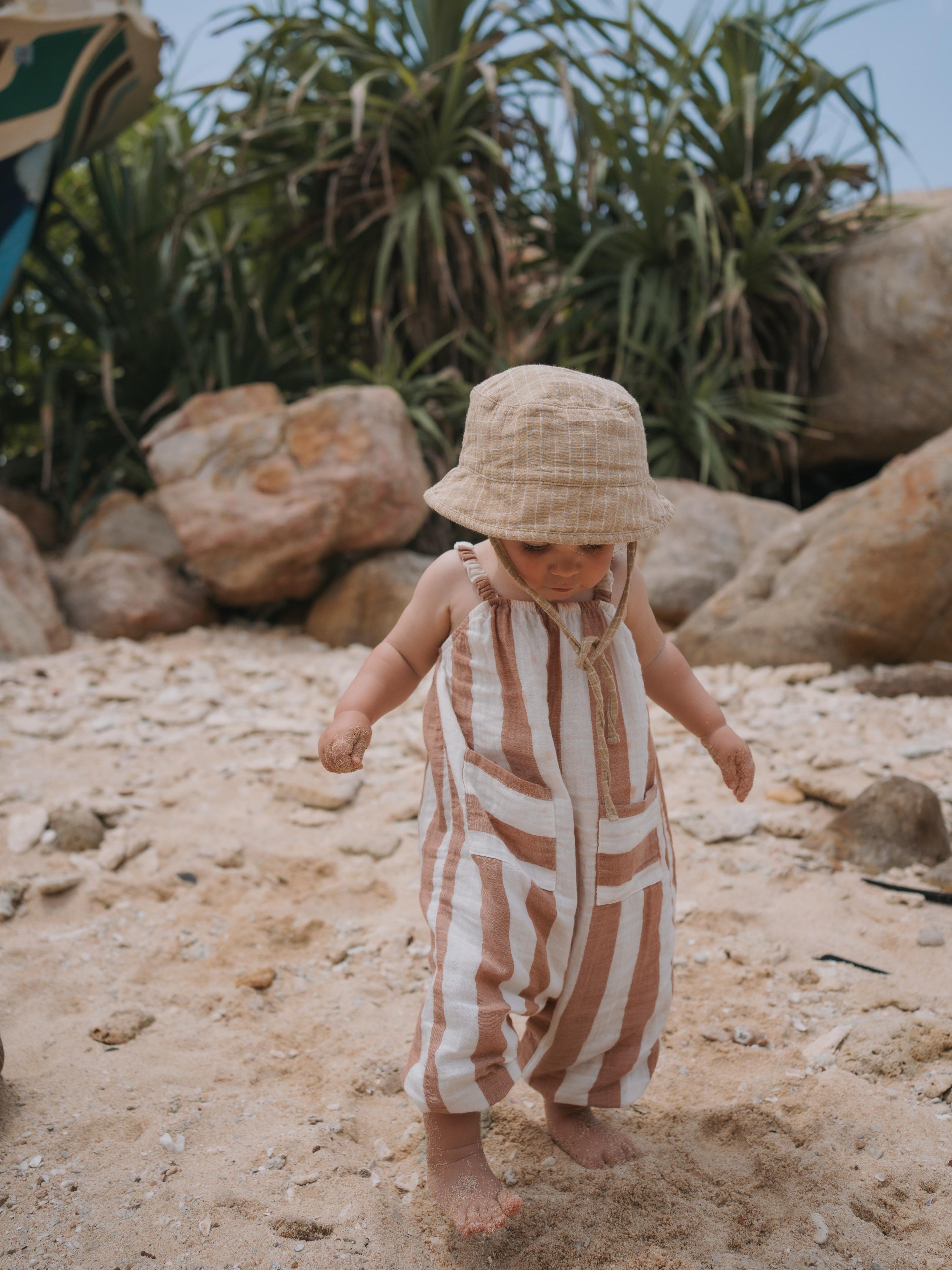 baby in sienna red striped muslin jumpsuit with gathered straps and musiln hat on the beach