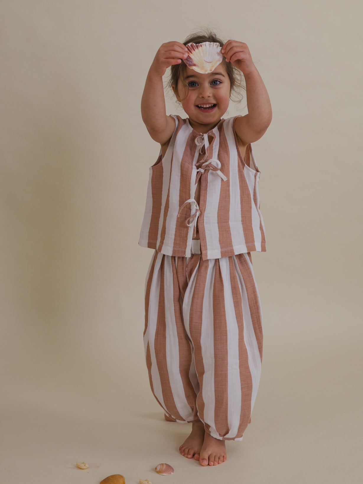 happy toddler wearing two way muslin ribbon front-tie resort top with red stripes holding seashell