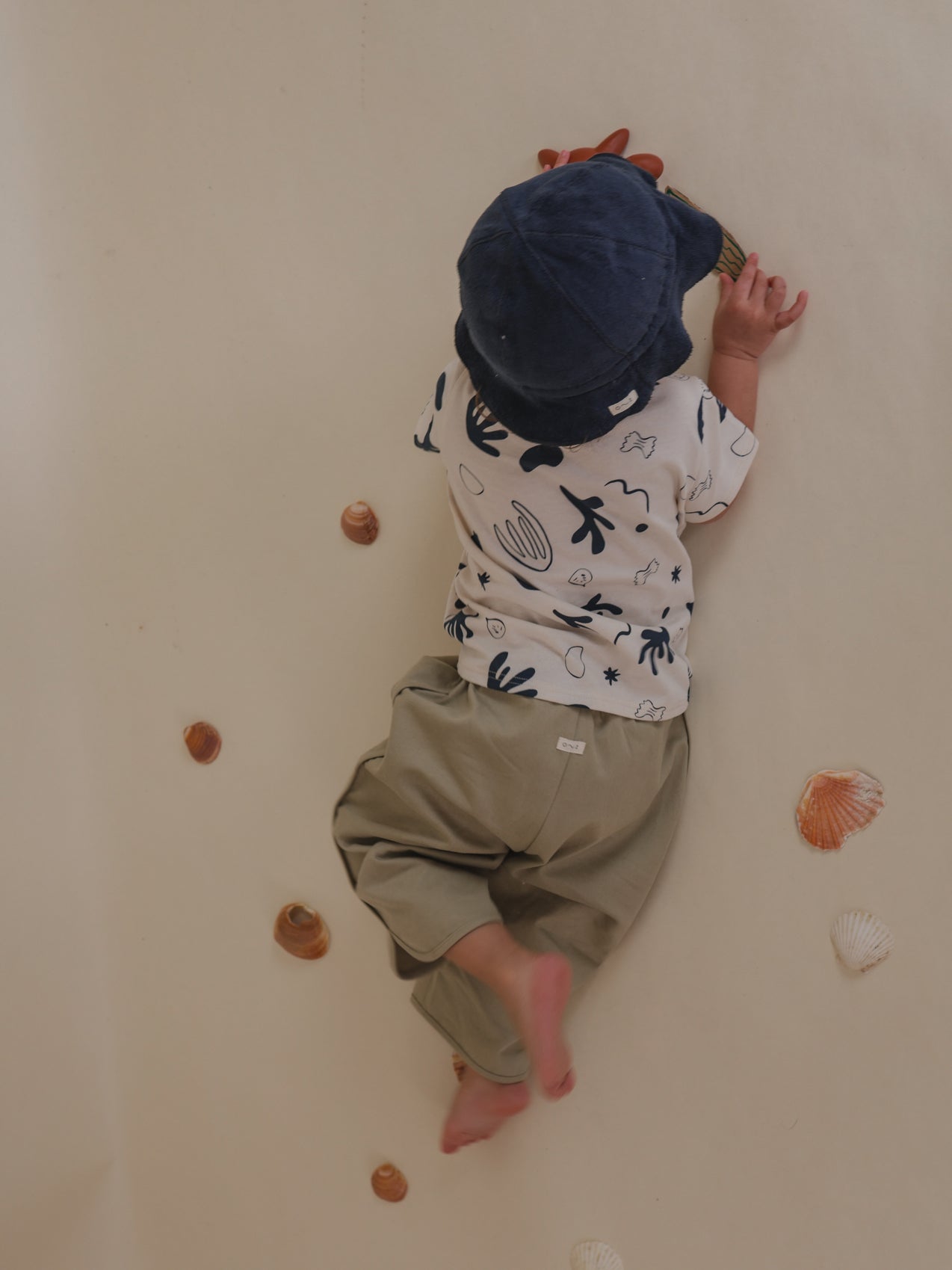 toddler in white undyed cotton classic capri t-shirt with ocean print lying on floor surrounded by seashells