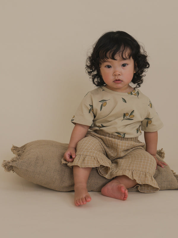 toddler wearing cream crew neck t-shirt with olives & grid frill culottes sitting on fringe cushion