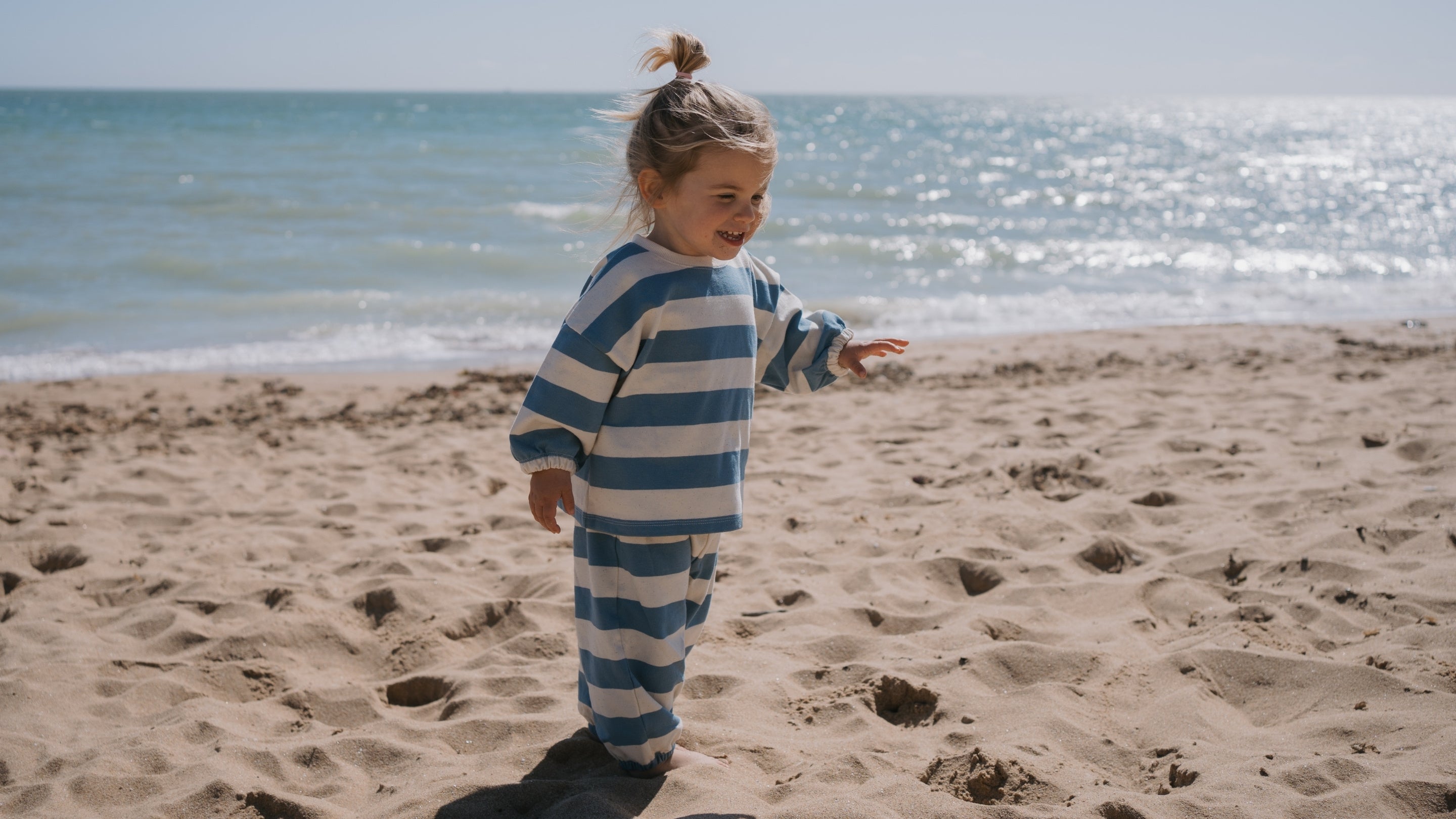 Child in blue and white striped outfit standing on a sandy beach with ocean in the background