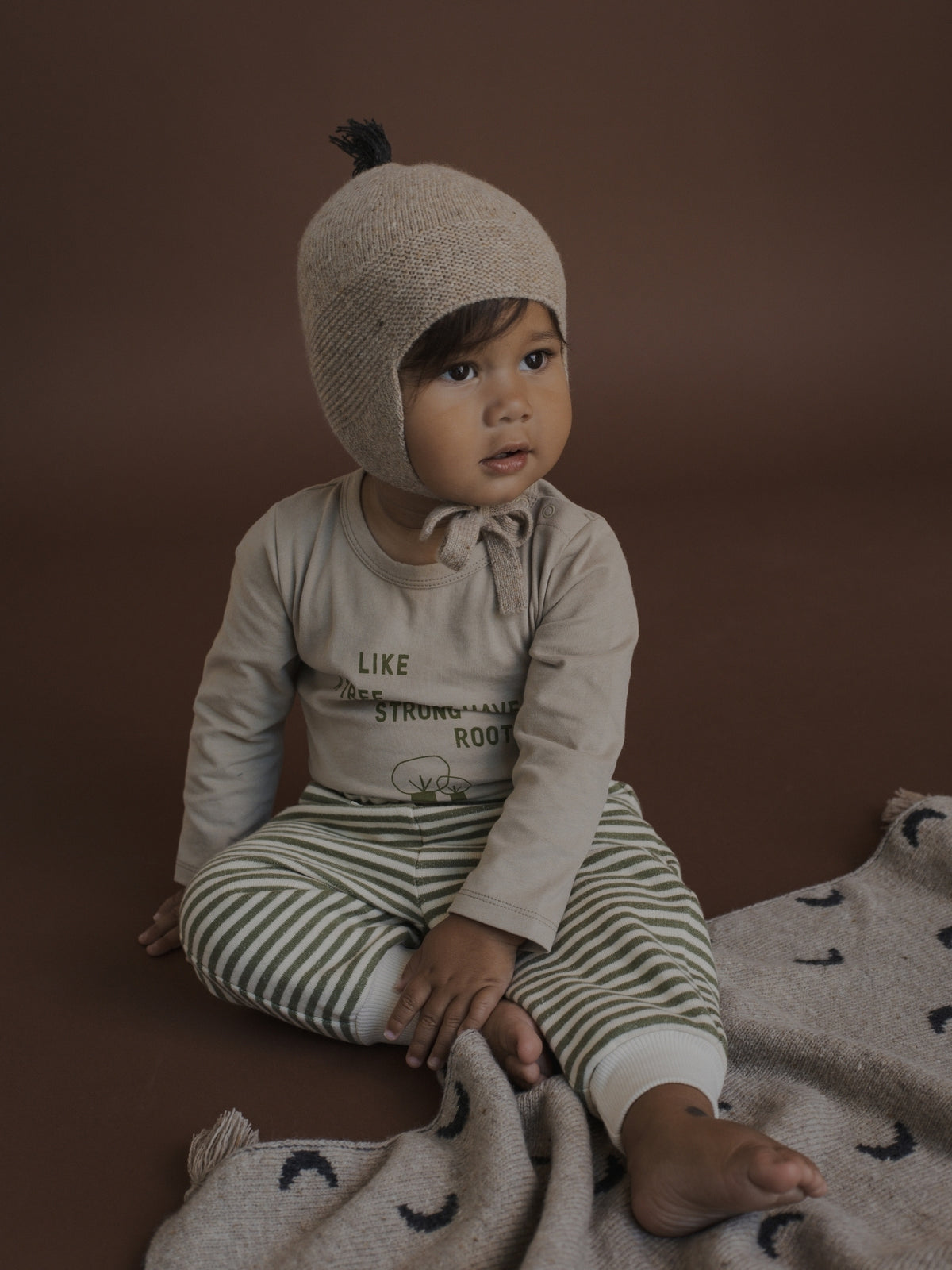 A baby sitting on a brown studio floor wearing a beige knit bonnet with a small black pom-pom, a beige long-sleeve shirt with text, and green and white striped pants, with a matching knit blanket.