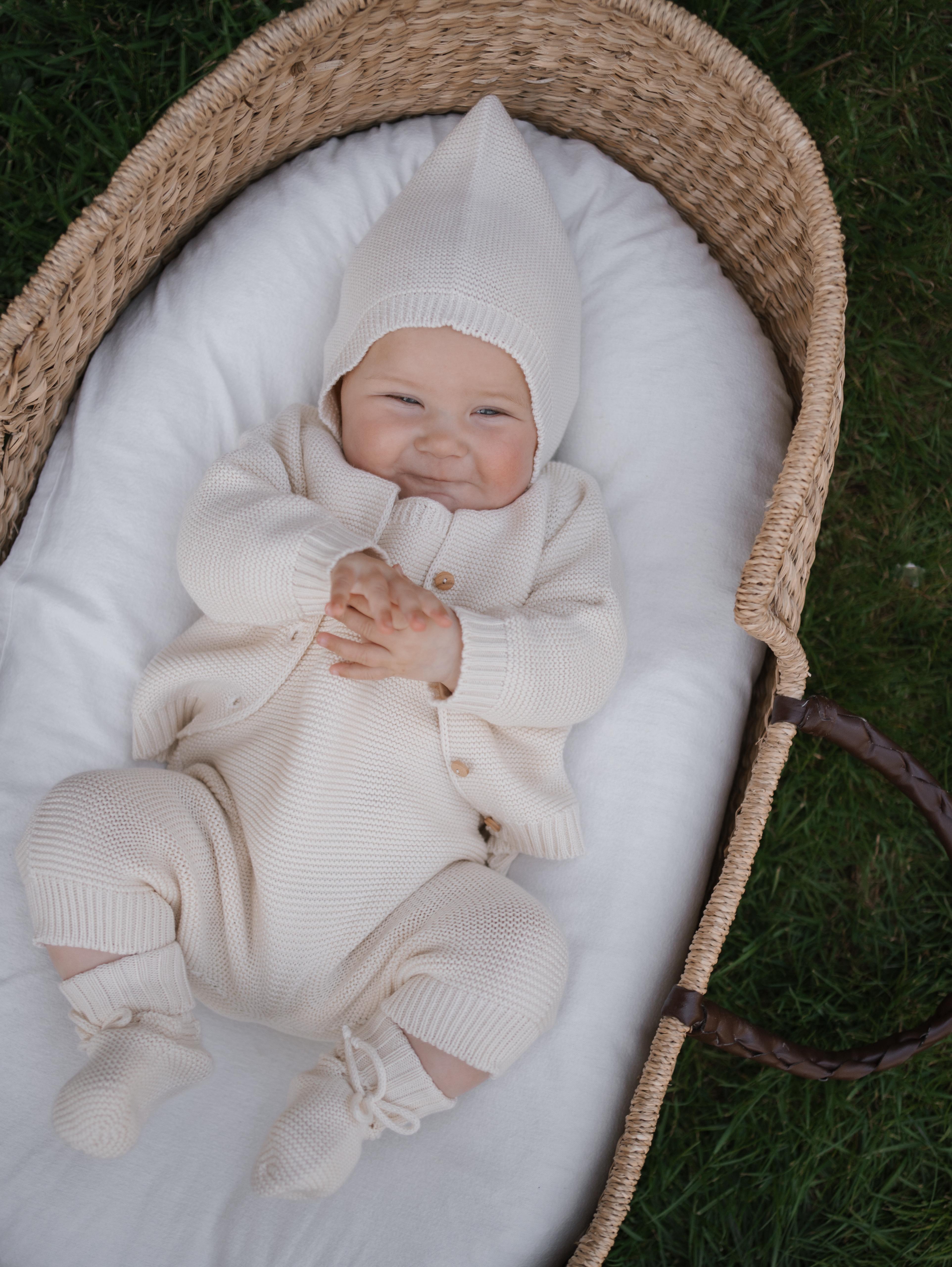happy baby wearing cream knitted cotton cardigan with matching hat, booties wiggling in moses basket