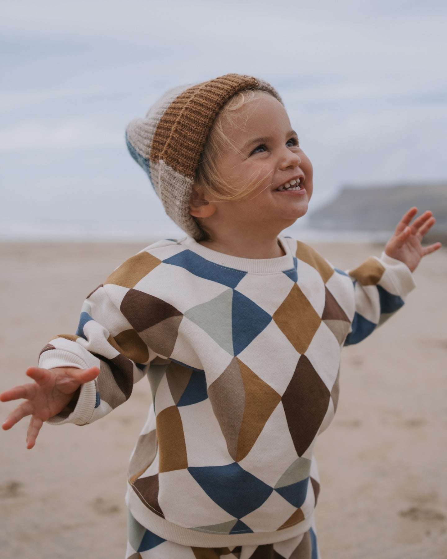 Child wearing a geometric-patterned organic cotton sweater and knit hat on a beach