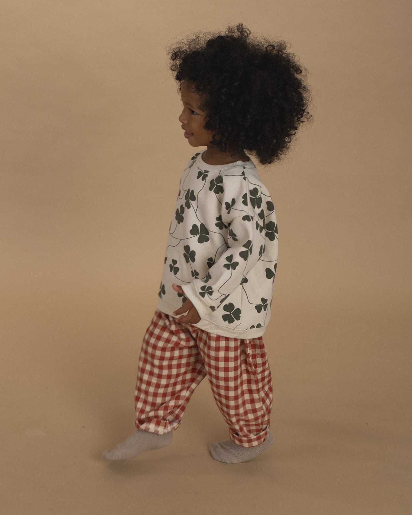 Child wearing an organic cotton white sweater with green leaf pattern and red checkered pants on a beige background