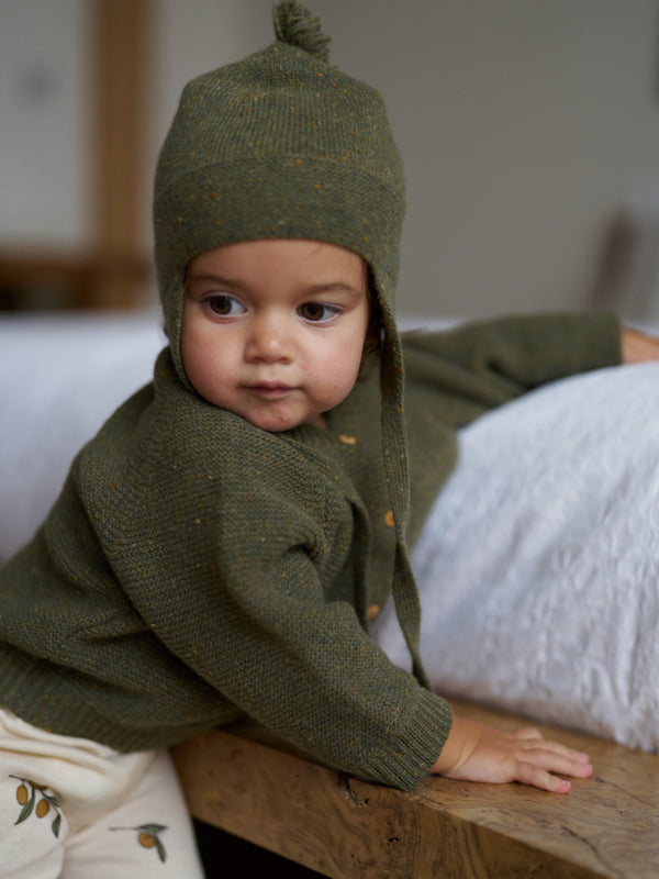 baby in forest moss green wool tassel hat with matching cardigan & cream leggings at bedside