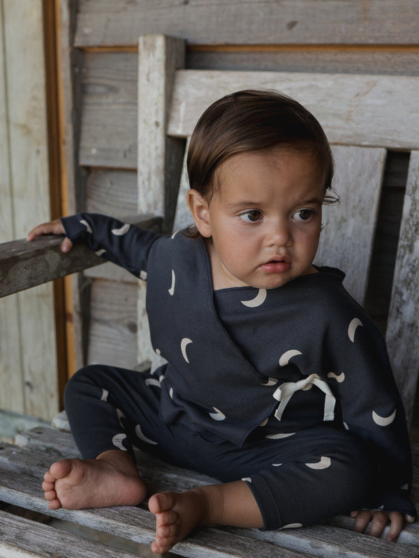 baby wearing black kimono with cream moons & white bow with matching leggings sitting on wooden bench