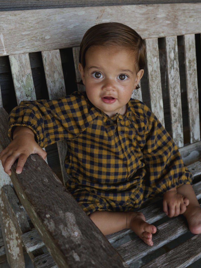 baby in flannel gingham onesie smiling into camera & sitting on wooden bench