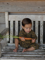 baby wearing navy & yellow checkered flannel onesie sitting on wooden bench holding a carrot