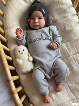 baby dressed in grey playsuit & black pixie bonnet with cream moons lying in rattan bassinet with toy