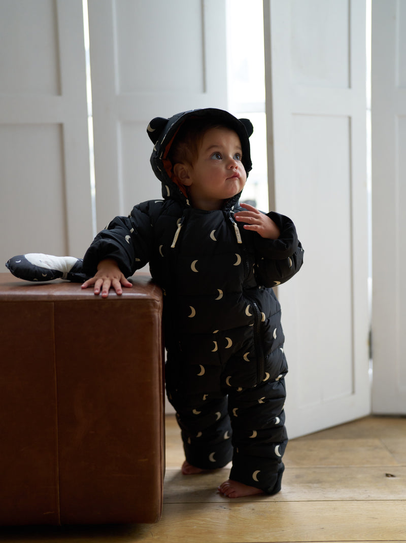 baby in black padded winter snowsuit with white moons & hood with ears standing beside ottoman