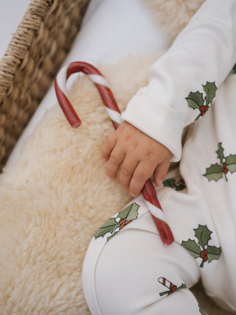 close up of baby in organic christmas holly & candy cane footed suit in cream holding candy cane