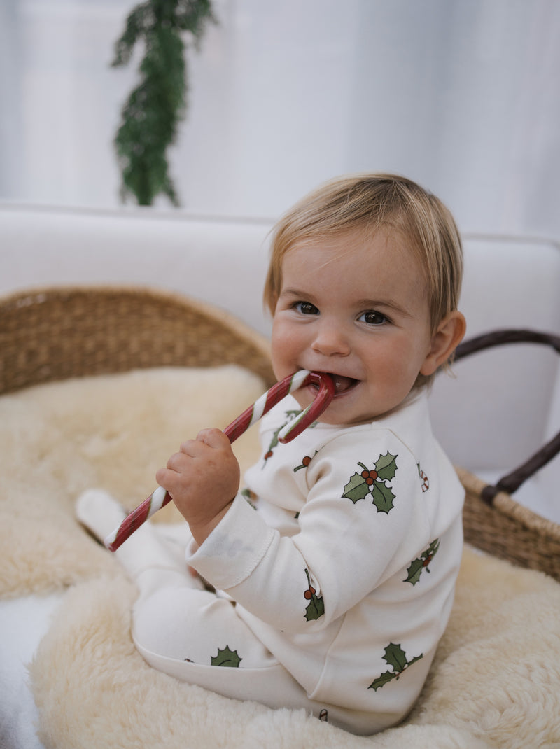 baby in cream organic footed suit with holly & candy cane print sitting in moses basket biting candy cane