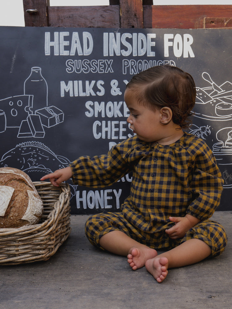 baby in navy & yellow checkered gingham flannel onesie sitting down pointing at bread in basket