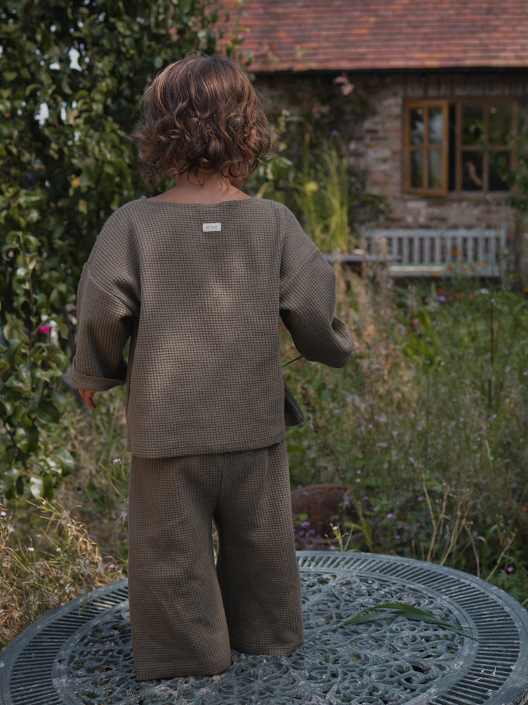 back view of child in olive waffle cotton sweatshirt & wide leg pants standing on patio table in garden