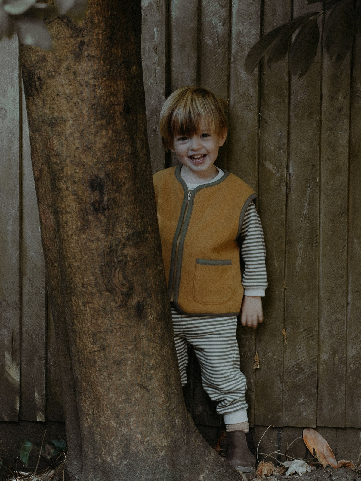 A young child with blond hair stands behind a large tree trunk in front of a weathered wooden fence, wearing a mustard yellow zip-front vest over a gray and white striped long-sleeve shirt and matching striped pants.