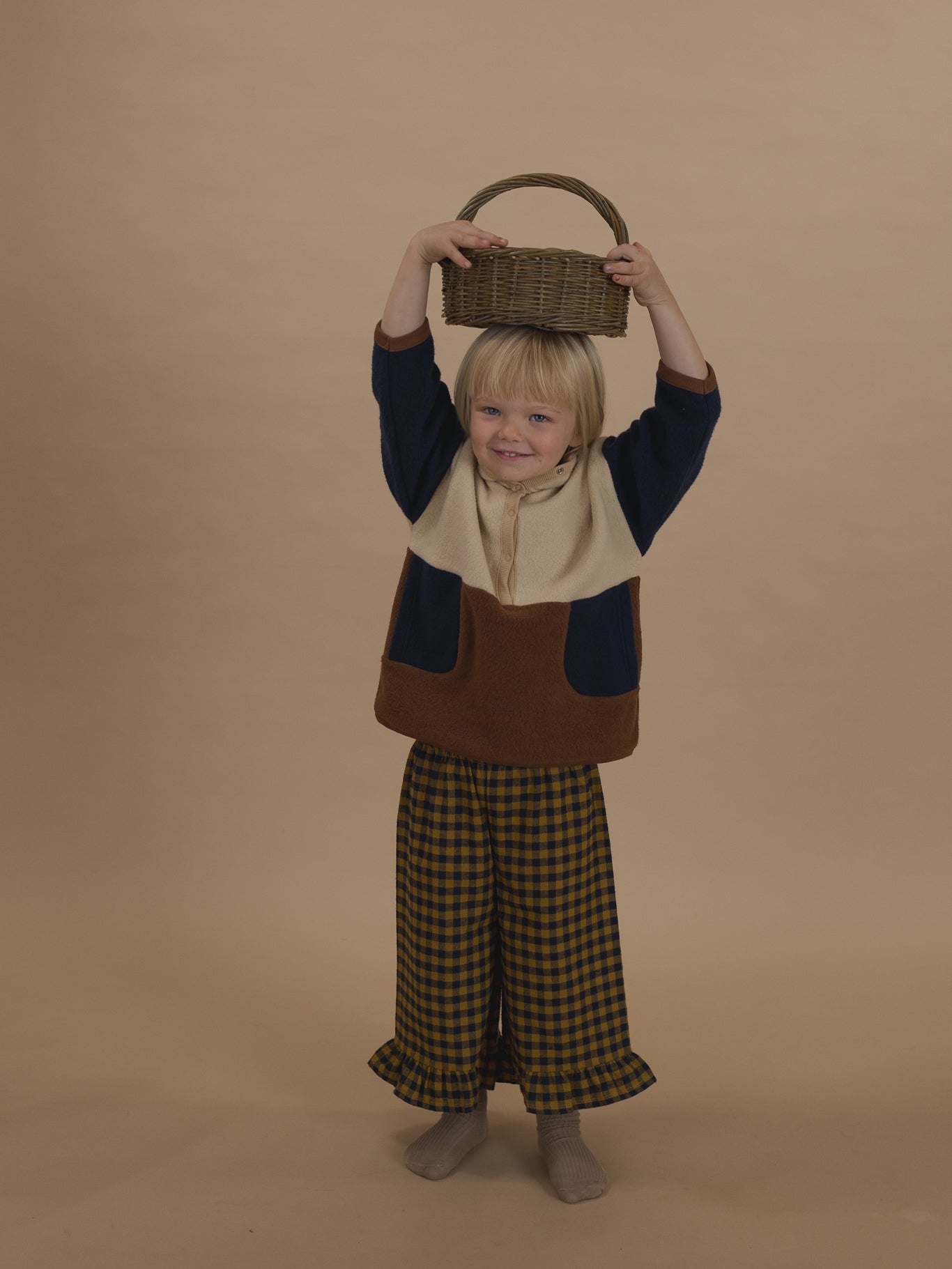 toddler in navy and yellow gingham frill culottes and colour block sweater, holding basket on top of head