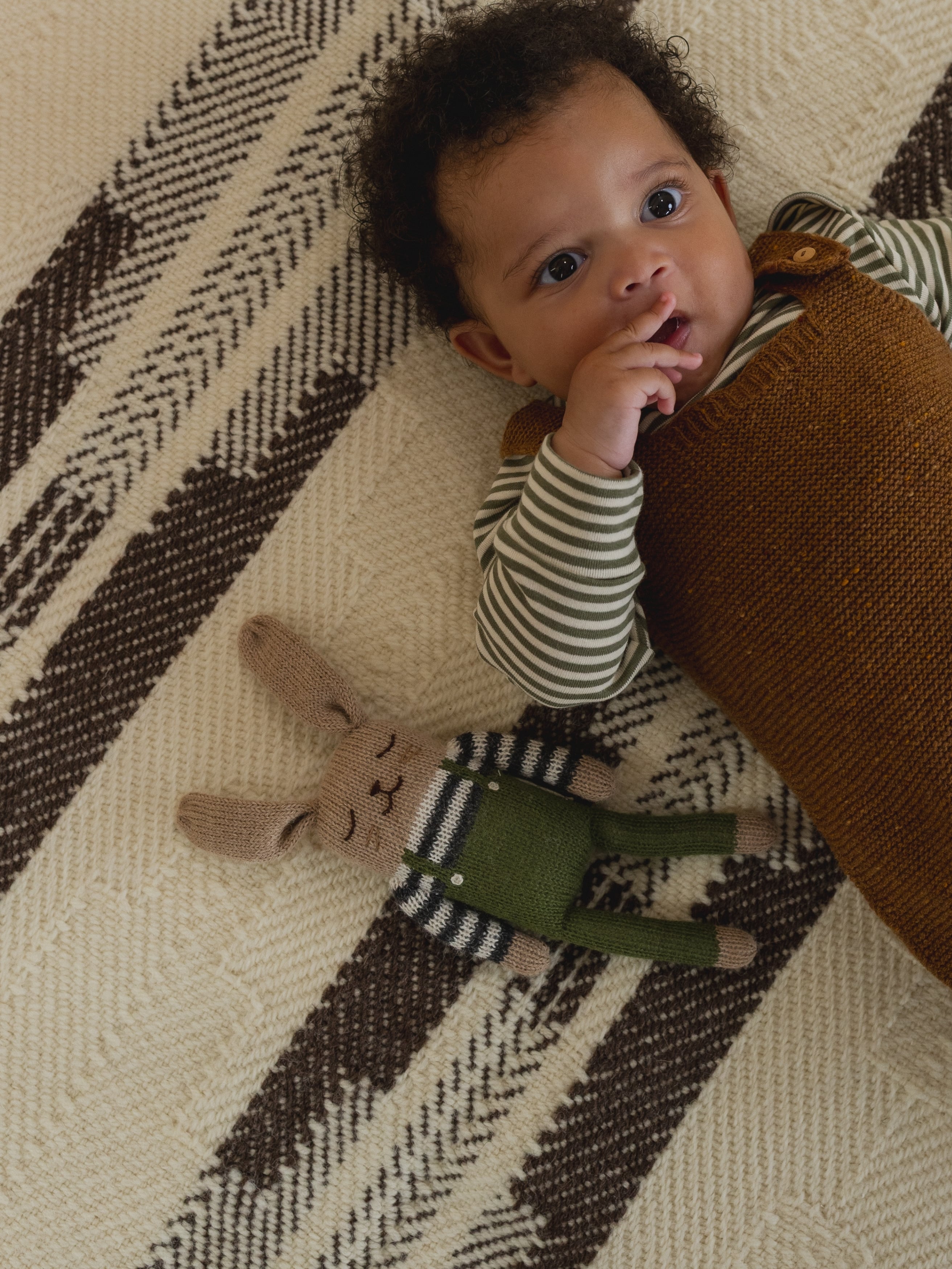 baby in salopette and top lying beside matching wool bunny toy in olive overalls with striped top