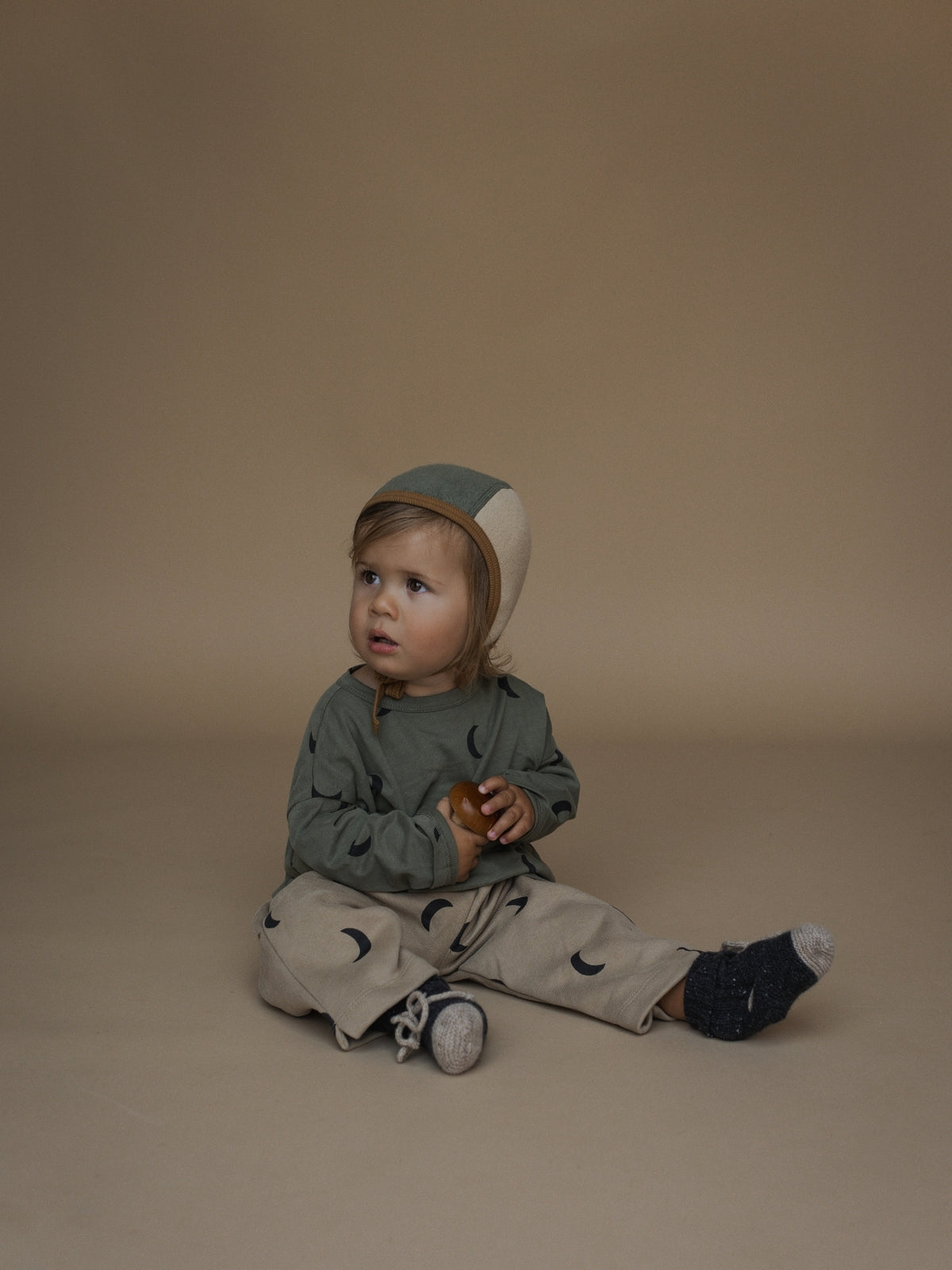 A toddler sitting on a warm beige studio floor wearing a muted sage green long-sleeve top, beige pants with black crescent moon prints, a matching green and beige bonnet, and dark socks.