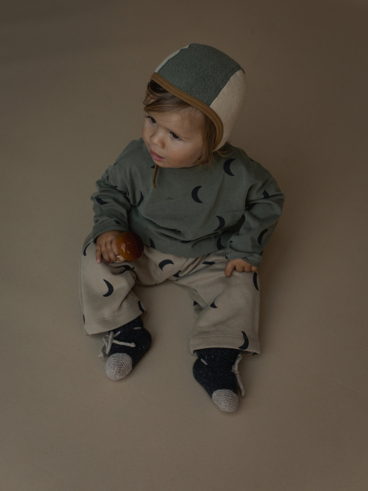 A baby sitting on a plain studio background wearing a muted sage green and beige hooded outfit with a crescent moon pattern, holding a small orange toy.
