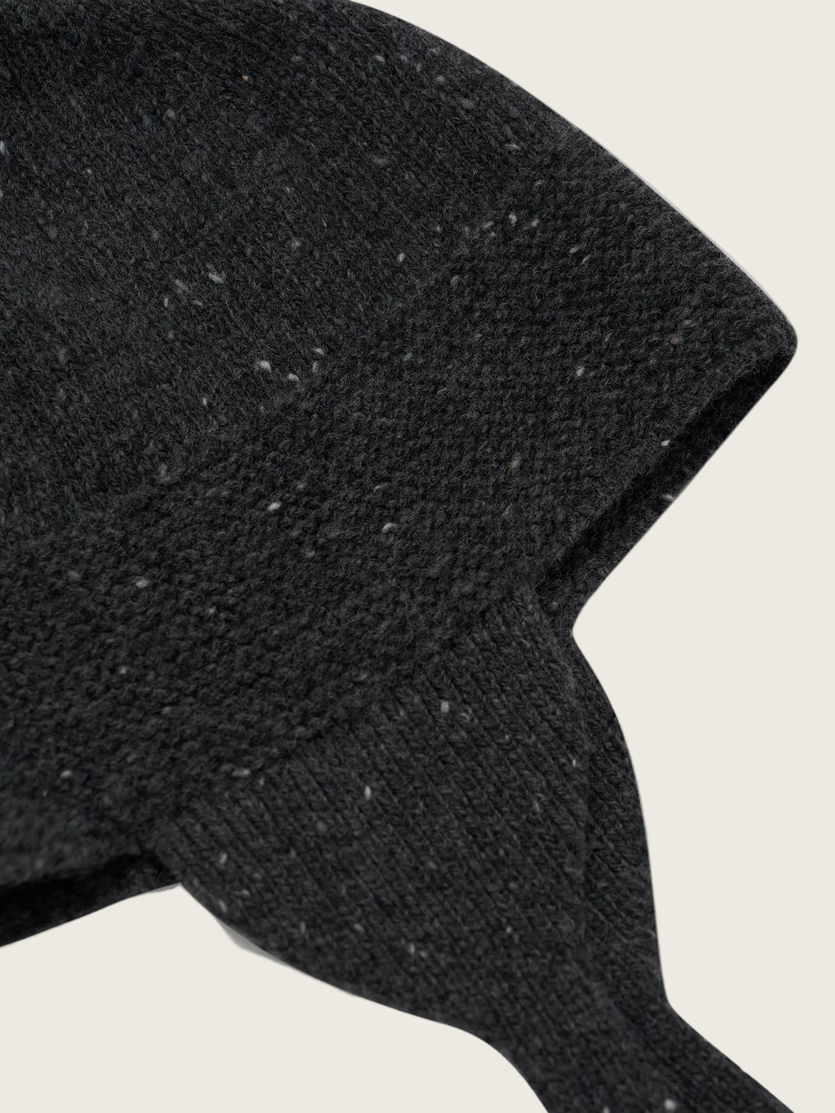 Close-up of a dark charcoal knit beanie with tiny white flecks, showing the textured fabric and ear flaps against a white background.