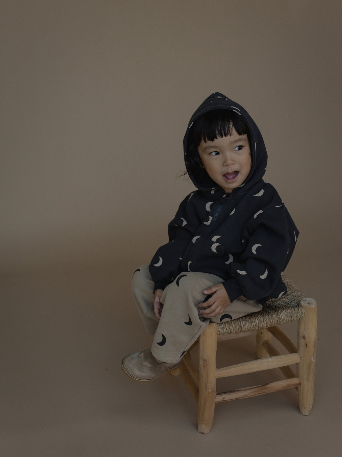 A toddler sits on a small wooden stool against a plain taupe studio background, wearing a dark navy hooded sweatshirt with a white crescent moon print and light beige pants.