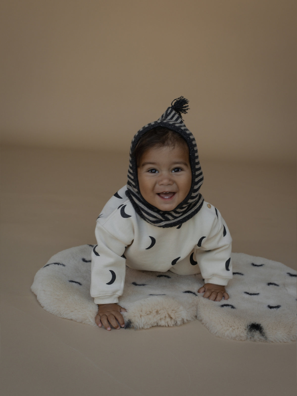 A baby smiling while crawling on a white sheepskin rug, wearing a cream sweatshirt with black crescent moon prints, a matching knit hood, and a striped knit neck warmer against a solid beige studio background.