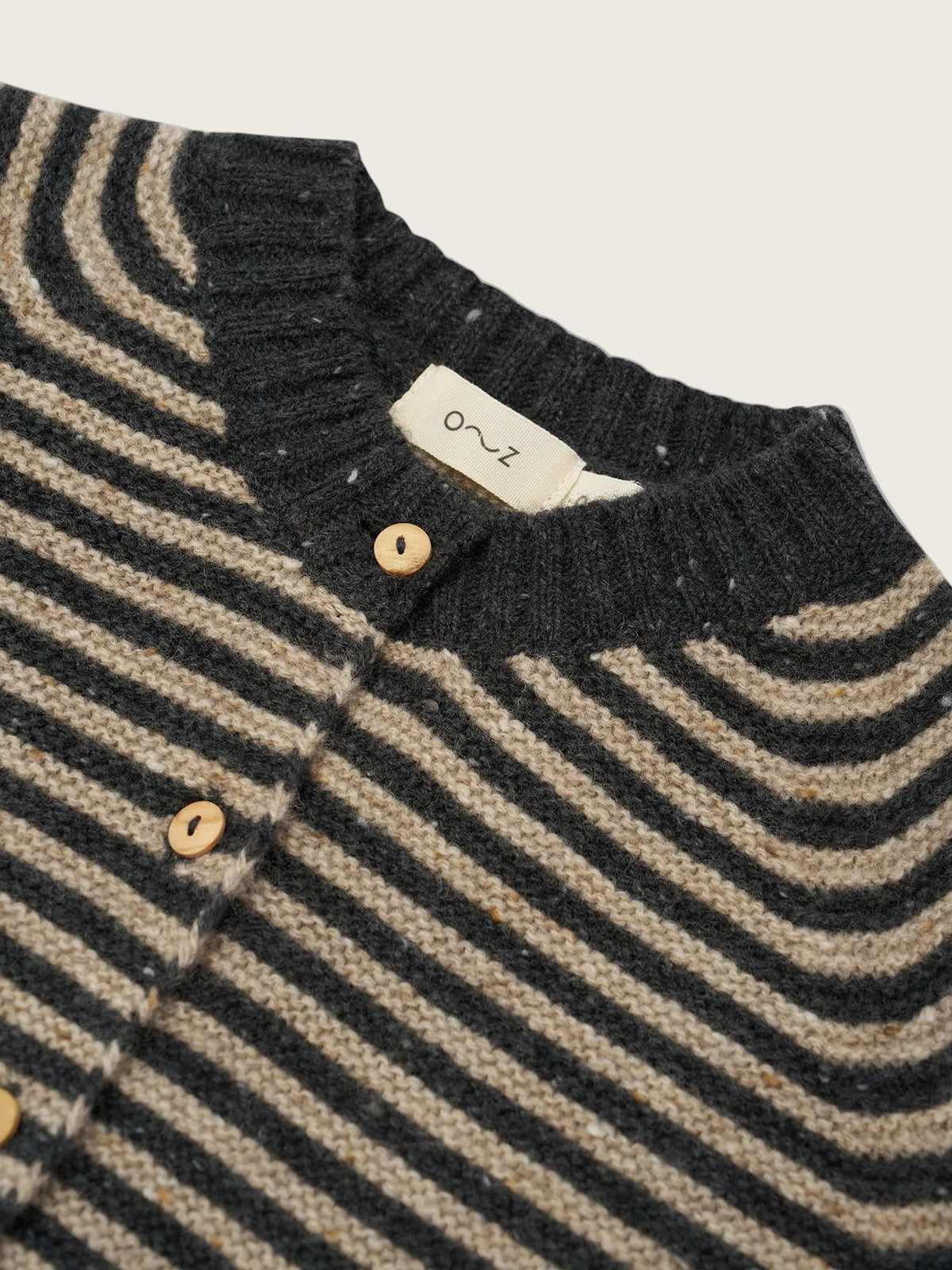 Close-up of a striped knit cardigan with dark charcoal and beige wavy horizontal stripes, featuring light wood buttons and a cream-colored neck label with the brand name 'oz'.