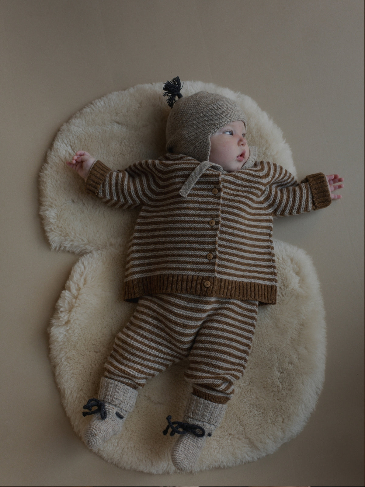 A baby lying on a cream sheepskin rug, wearing a matching knit three-piece outfit with brown and beige horizontal stripes, a beige bonnet, and knit booties.