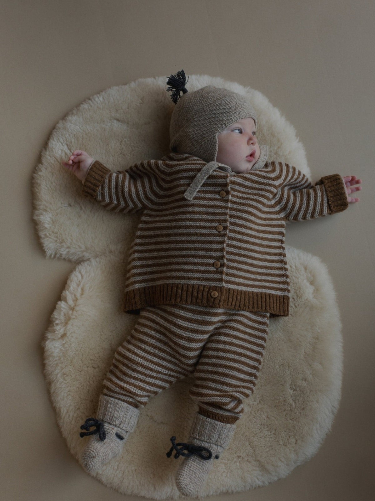A baby lying on a cream sheepskin rug, wearing a matching knit three-piece outfit with brown and beige horizontal stripes, a beige bonnet, and knit booties.
