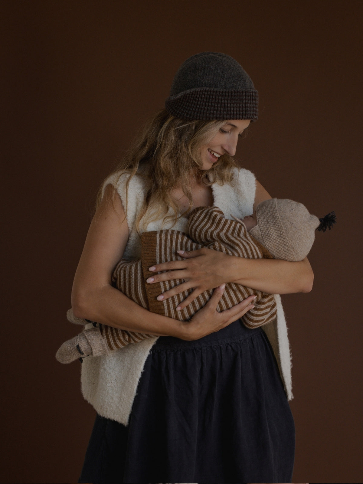 A woman wearing a dark gray checkered beanie, a cream sherpa vest, and a dark navy dress holds a baby wrapped in a brown and beige striped knit blanket against a solid dark brown background.