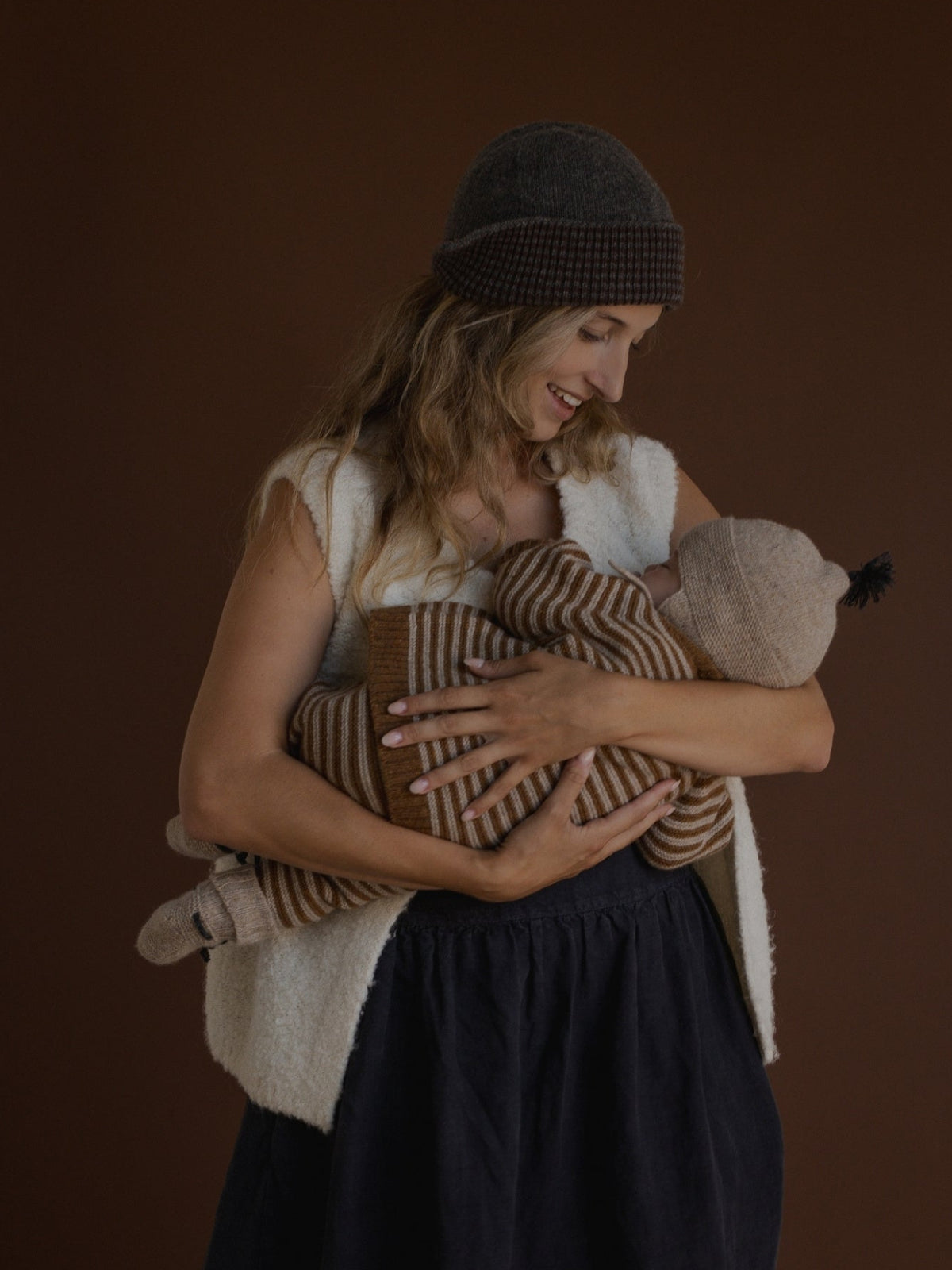A woman wearing a dark gray checkered beanie, a cream sherpa vest, and a dark navy dress holds a baby wrapped in a brown and beige striped knit blanket against a solid dark brown background.
