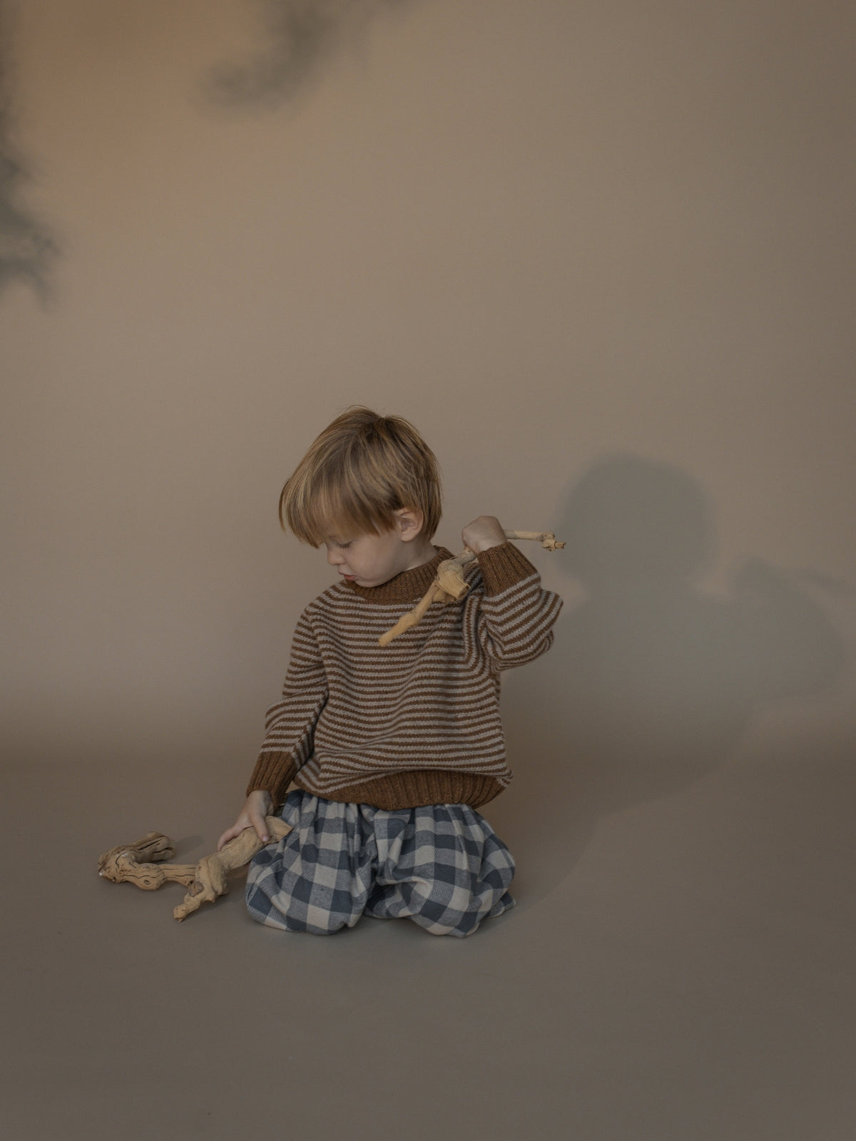A young child kneeling on a warm beige studio floor, wearing a brown and beige striped knit sweater and blue-and-white gingham pants, holding a wooden toy.