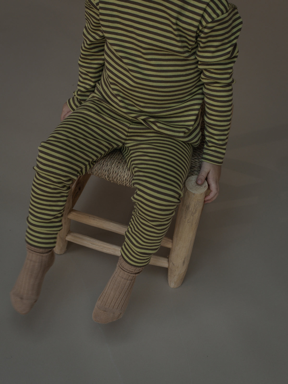 A child wearing a matching two-piece set with olive green and dark brown horizontal stripes, sitting on a small wooden stool against a neutral gray background.