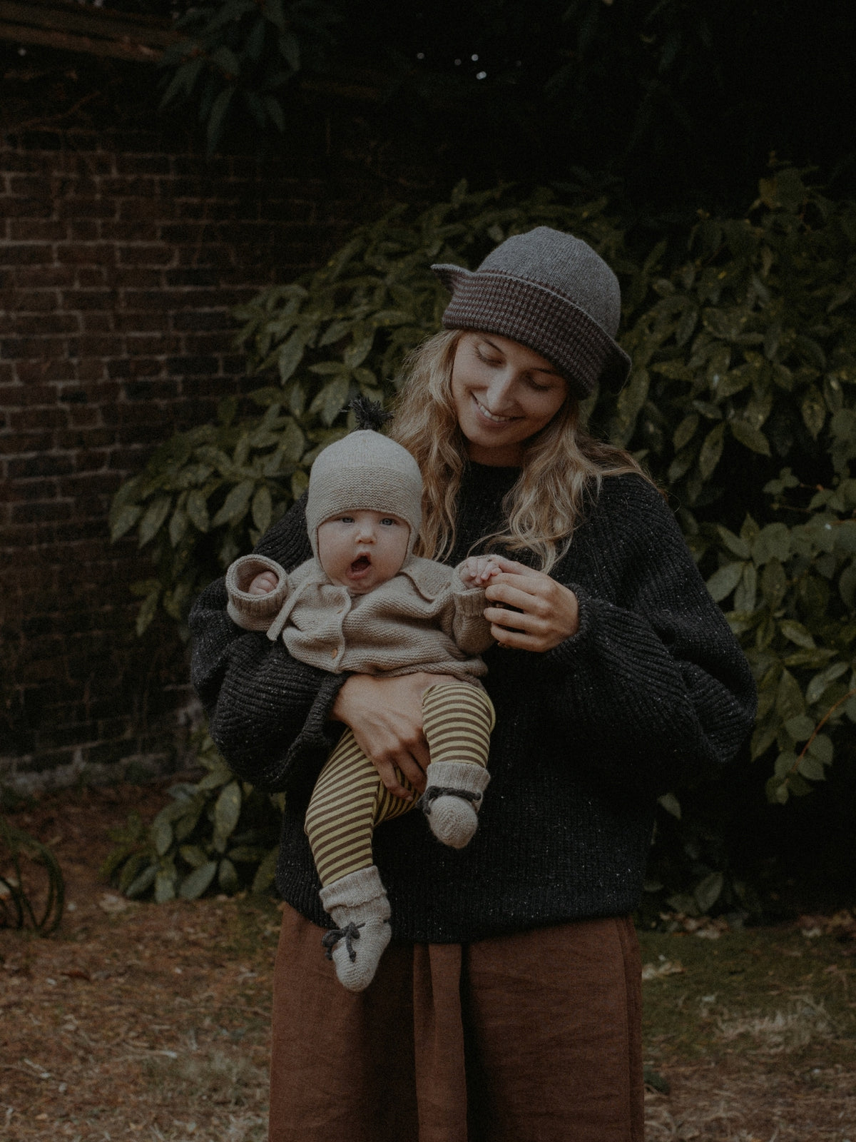 A woman in a dark knit sweater and gray beanie holds a baby wearing a light knit beanie, cream top, and striped leggings in an outdoor garden setting.