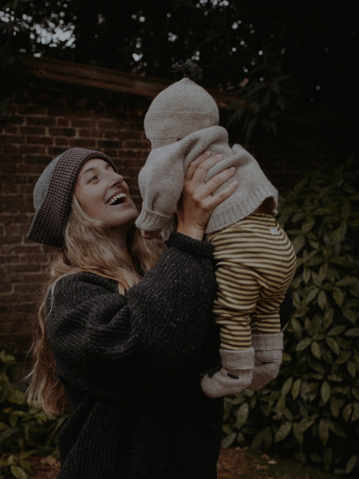 A woman in a dark knit sweater and checkered beanie holds a baby wearing a light beige knit bonnet and yellow-and-brown striped pants, set against a dark outdoor background with a brick wall and greenery.
