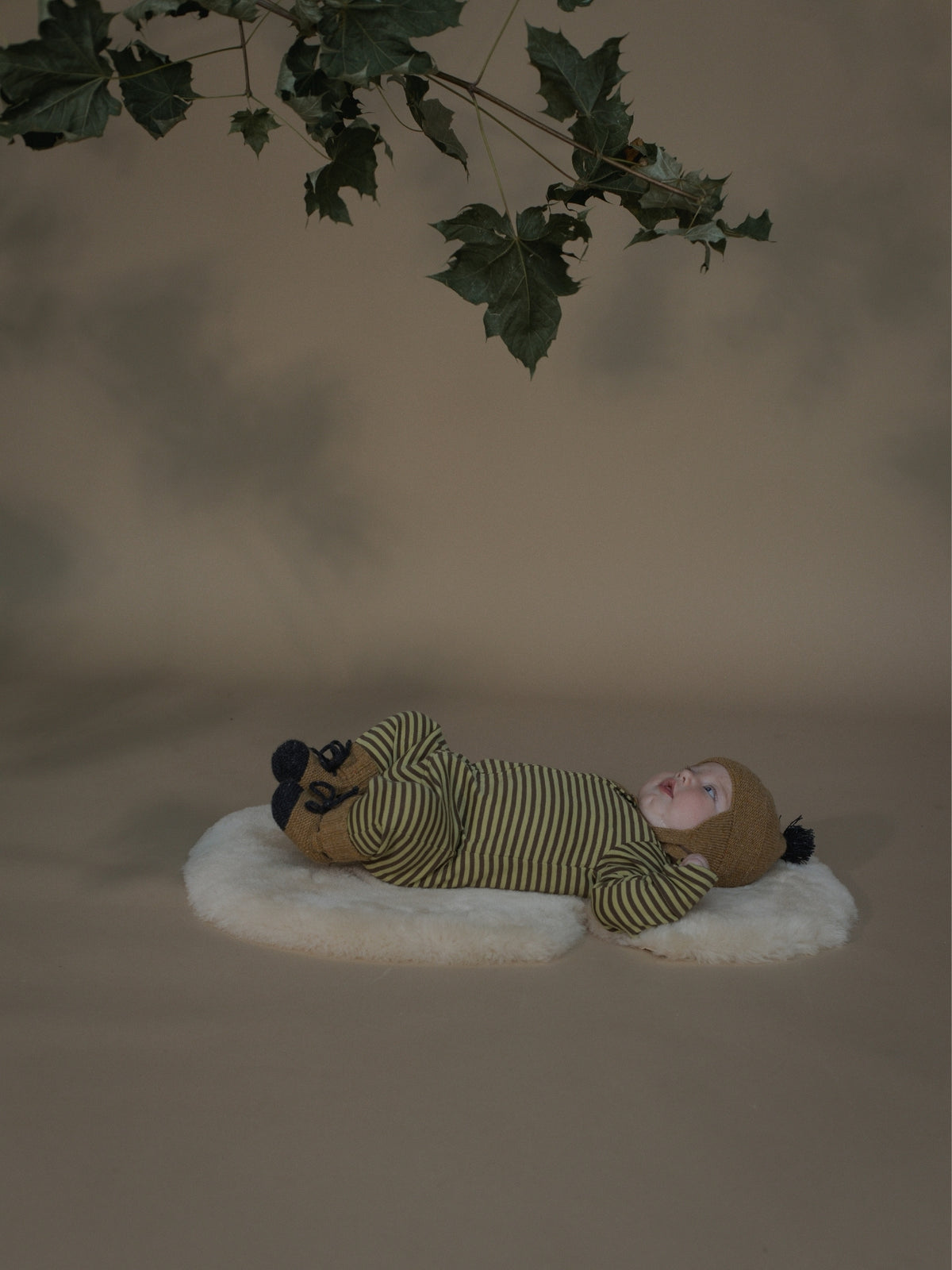A baby lying on a white faux-fur cushion, wearing a brown knit beanie and a green and beige striped one-piece outfit, posed under hanging green leaves against a warm beige studio backdrop.