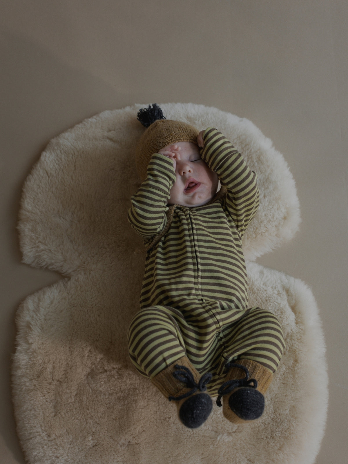 A baby lying on a cream sheepskin rug, wearing a green and olive striped footed one-piece with a tan hat and dark booties.