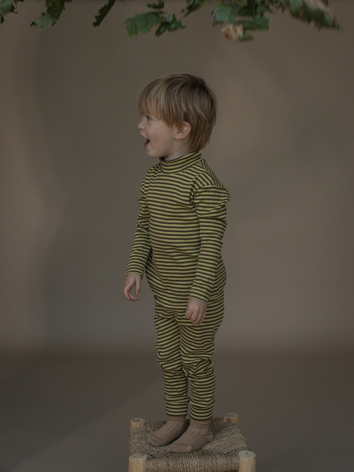 A toddler stands in profile wearing a matching olive green and mustard yellow striped long-sleeve turtleneck top and striped pants, posed on a small woven stool against a warm taupe studio backdrop.