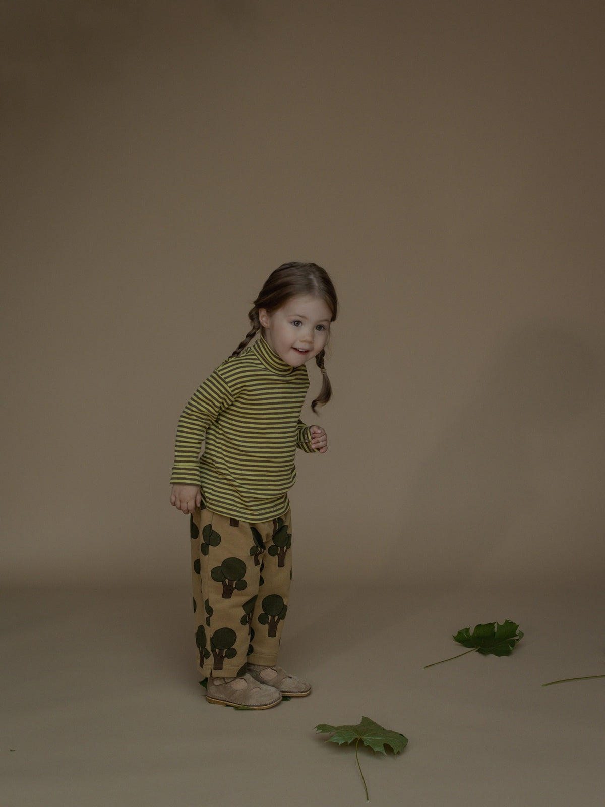 A young girl with braided hair stands in a studio wearing a green and yellow striped turtleneck top and tan pants with a dark green mushroom print. She is posed against a solid warm brown backdrop with two green leaves on the floor.