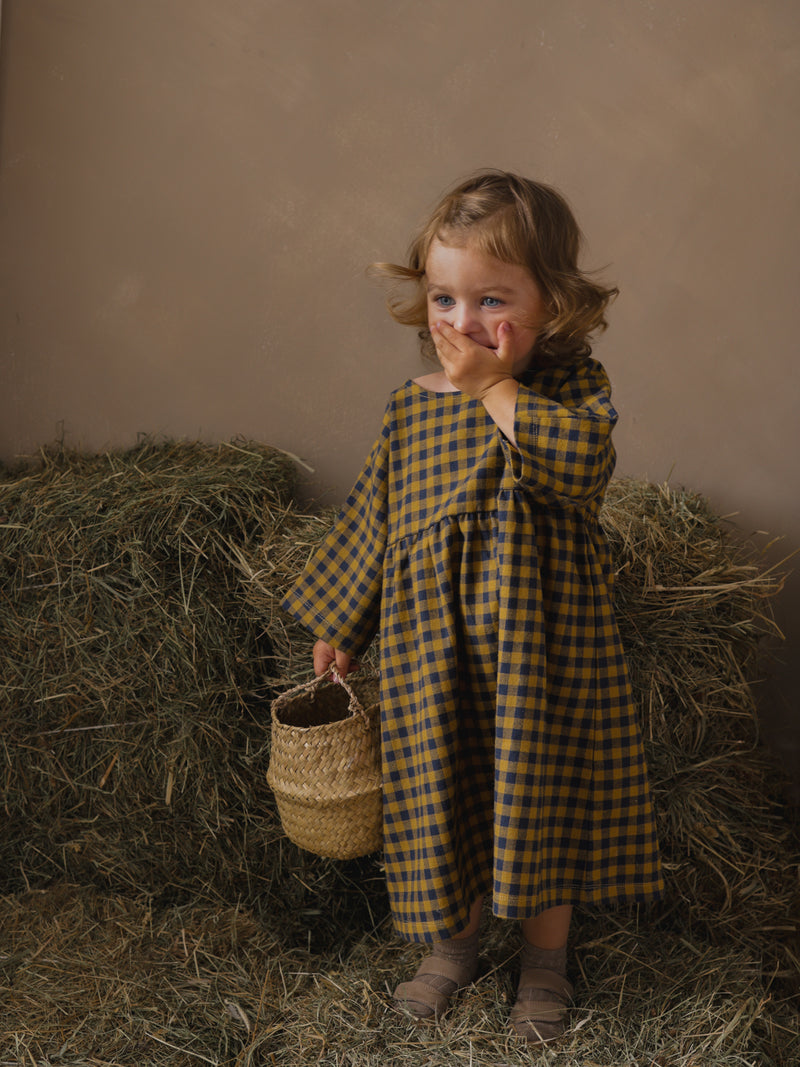 child holding basket giggling with hand over mouth wearing navy & yellow gingham dress brown sandals & socks