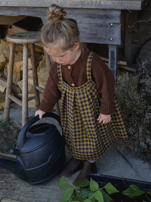 child in brown wool cardigan & gingham apron-style skirt holding watering can