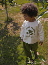 child in cream sweatshirt with FARMERS MARKET ORGANIC ZOO logo & green long pants barefoot in garden