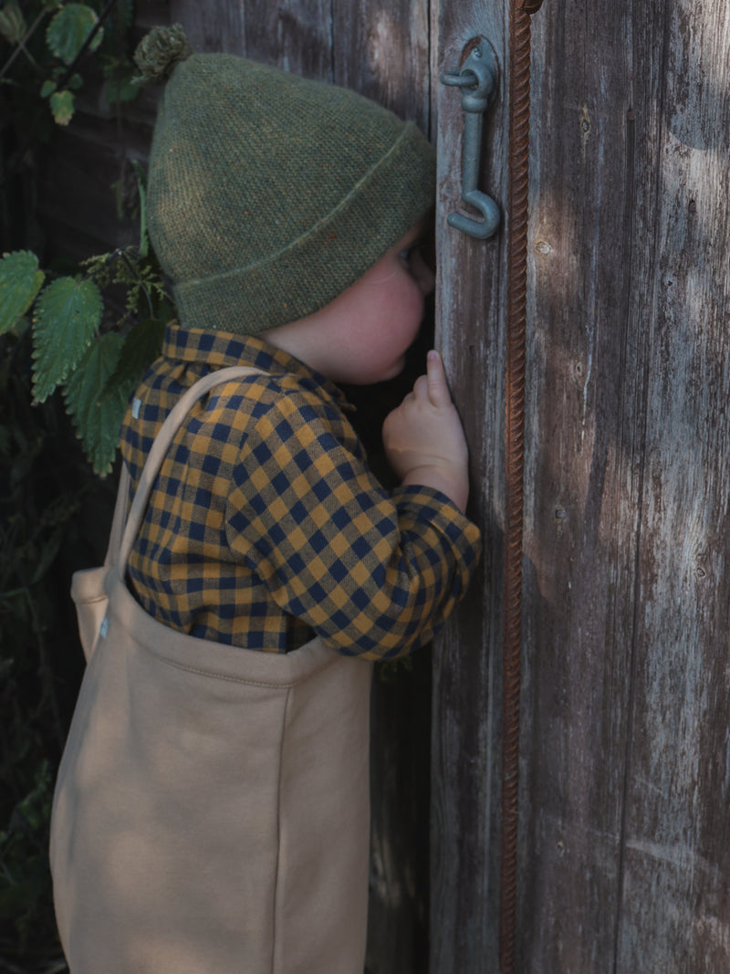 child in gingham flannel shirt & terry cotton beige overalls & green beanie peeking through wooden door