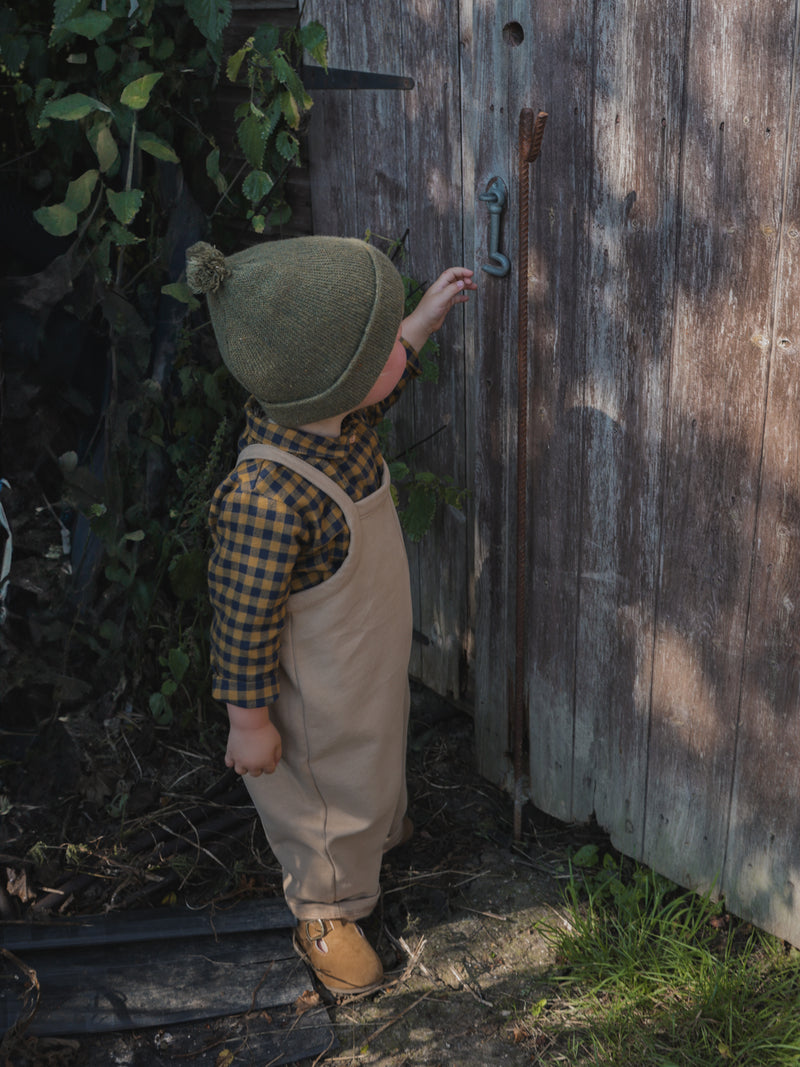 child in gingham flannel shirt terry cotton & beige overalls & brown shoes reaching for wooden door in garden